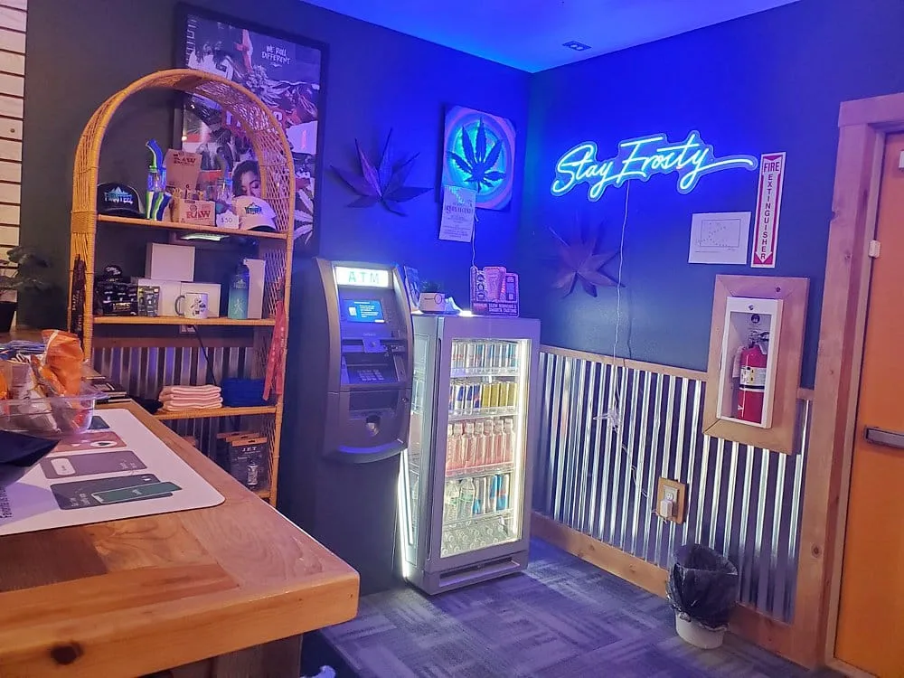 Neon-lit cafe corner with a Stay Frosty sign, blue wall, ATM, and a drink vending refrigerator.