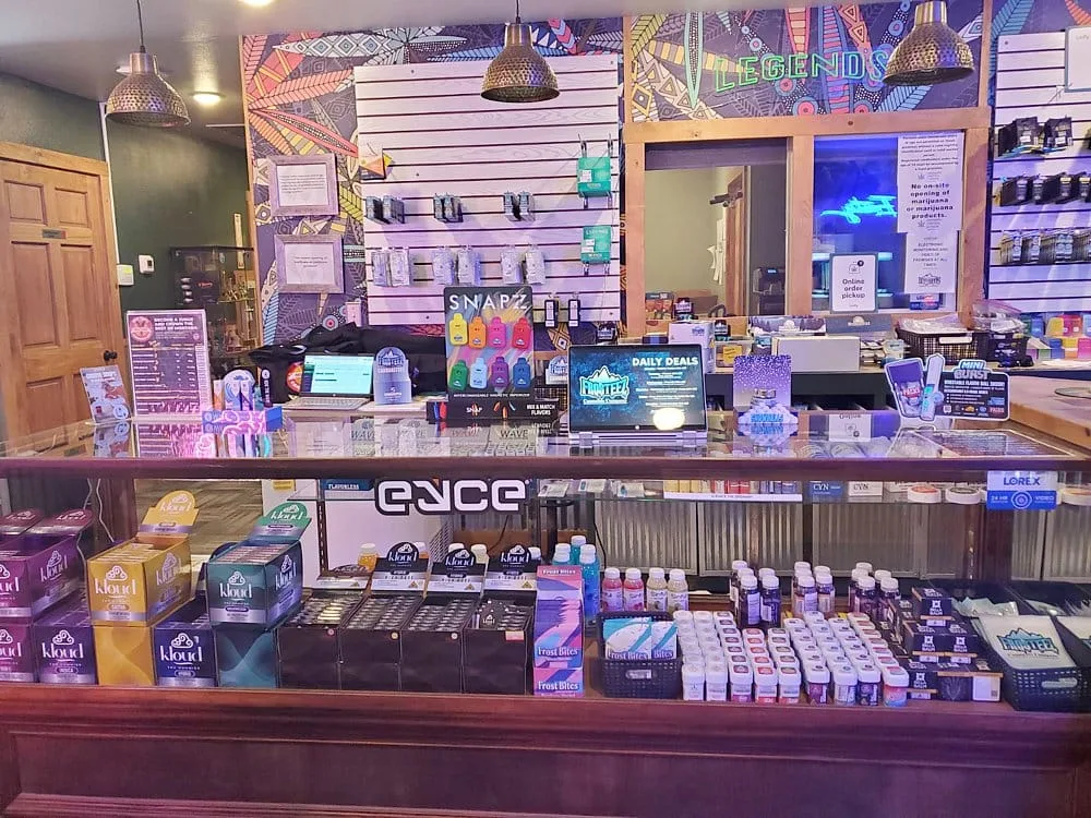 Colorful storefront counter with glass display of products, neon 'LEGENDS' sign, and single laptop on the counter; shelves and wall display behind.