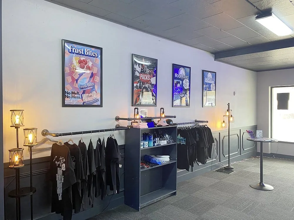 Interior of a clothing shop with black t-shirts on a wall rack, a central navy shelf with folded clothes and accessories, and framed posters on the wall under soft lighting.